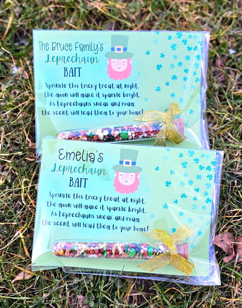 May include: Two clear plastic bags with green and white patterned paper inside. Each bag contains a small glass vial filled with colorful sprinkles and a gold ribbon. The paper inside each bag has a leprechaun illustration and the text "The Bruce Family's Leprechaun Bait" and "Emelia's Leprechaun Bait". The text reads: "Sprinkle this tricky treat at night. The moon will make it sparkle bright. As Leprechauns sneak and roam. The scent will lead them to your home!"