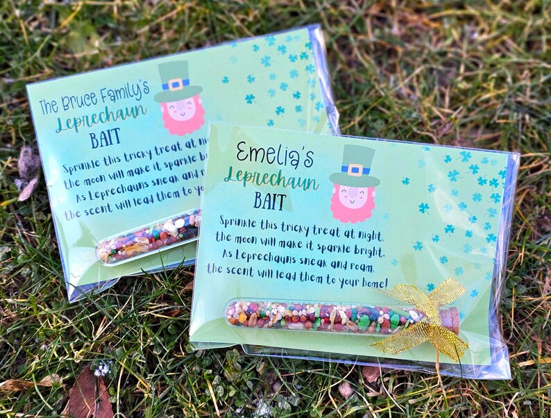 May include: Two clear plastic bags with green and white cards inside. The cards have a leprechaun illustration and the text "The Bruce Family's Leprechaun Bait" and "Emelia's Leprechaun Bait". Each bag contains a small glass vial filled with colorful sprinkles.