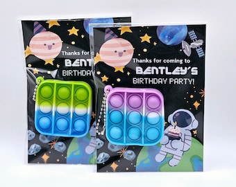 May include: Two party favor bags with a space theme. Each bag has a pop it keychain attached. The pop it keychains are blue, green, and purple. The bags have the text "Thanks for coming to Bentley's Birthday Party!" printed on them.