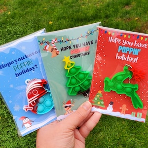 May include: Three Christmas-themed cards with the text "Hope you have a POPPIN' holiday?" and "HOPE YOU HAVE A POPPIN' CHRISTMAS!" and "Hope you have a POPPIN' holiday!" Each card features a different pop-it toy in the shape of a snowman, a Christmas tree, and a reindeer.
