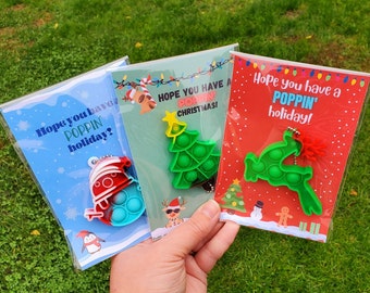 May include: Three Christmas-themed cards with the text "Hope you have a POPPIN' holiday?" and "HOPE YOU HAVE A POPPIN' CHRISTMAS!" and "Hope you have a POPPIN' holiday!" Each card features a different pop-it toy in the shape of a snowman, a Christmas tree, and a reindeer.
