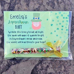May include: A green card with the text "Emelia's Leprechaun Bait" and a cartoon leprechaun. The card is in a clear plastic bag with a gold ribbon and a small glass tube filled with colorful sprinkles.