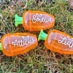 May include: Three orange carrot-shaped pencil holders with green tops. The holders are personalized with the names "Emeliq", "Atticus", and "Liam".