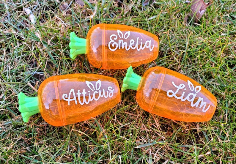 May include: Three orange carrot-shaped pencil holders with green tops. The holders are personalized with the names "Emeliq", "Atticus", and "Liam".