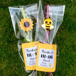 May include: Two clear plastic bags with yellow and pink straws, each with a bee or sunflower charm. The bags have yellow tags with the text "Thanks for BEE-IN here! From: Emelia" and "Thanks for BEE-ING here! From: Emelia".