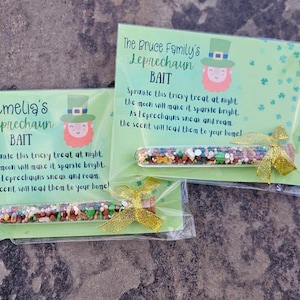 May include: Two green and white cards with instructions for a leprechaun bait. The cards have a picture of a leprechaun on them. The cards are in clear plastic bags with a small glass tube filled with colorful sprinkles. The tube is tied with a gold ribbon.