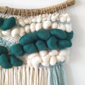 May include: A woven wall hanging with a natural wood dowel at the top. The hanging features a variety of textures and colors, including white, green, and blue. The bottom of the hanging has long white fringe and a smaller blue fringe.
