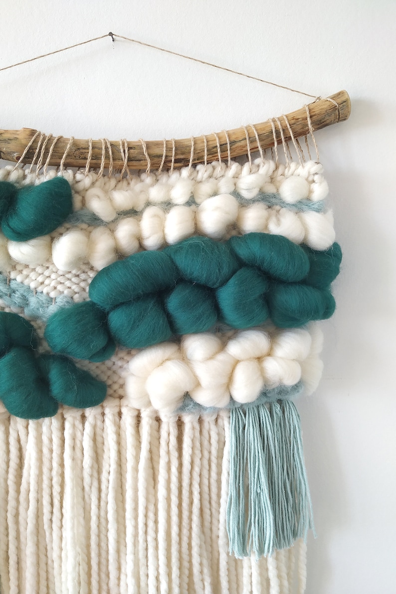 May include: A woven wall hanging with a natural wood dowel at the top. The hanging features a variety of textures and colors, including white, green, and blue. The bottom of the hanging has long white fringe and a smaller blue fringe.