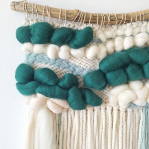 May include: A woven wall hanging with a driftwood frame. The hanging features a green and white color scheme with a variety of textures. The hanging has a fringe of white yarn.