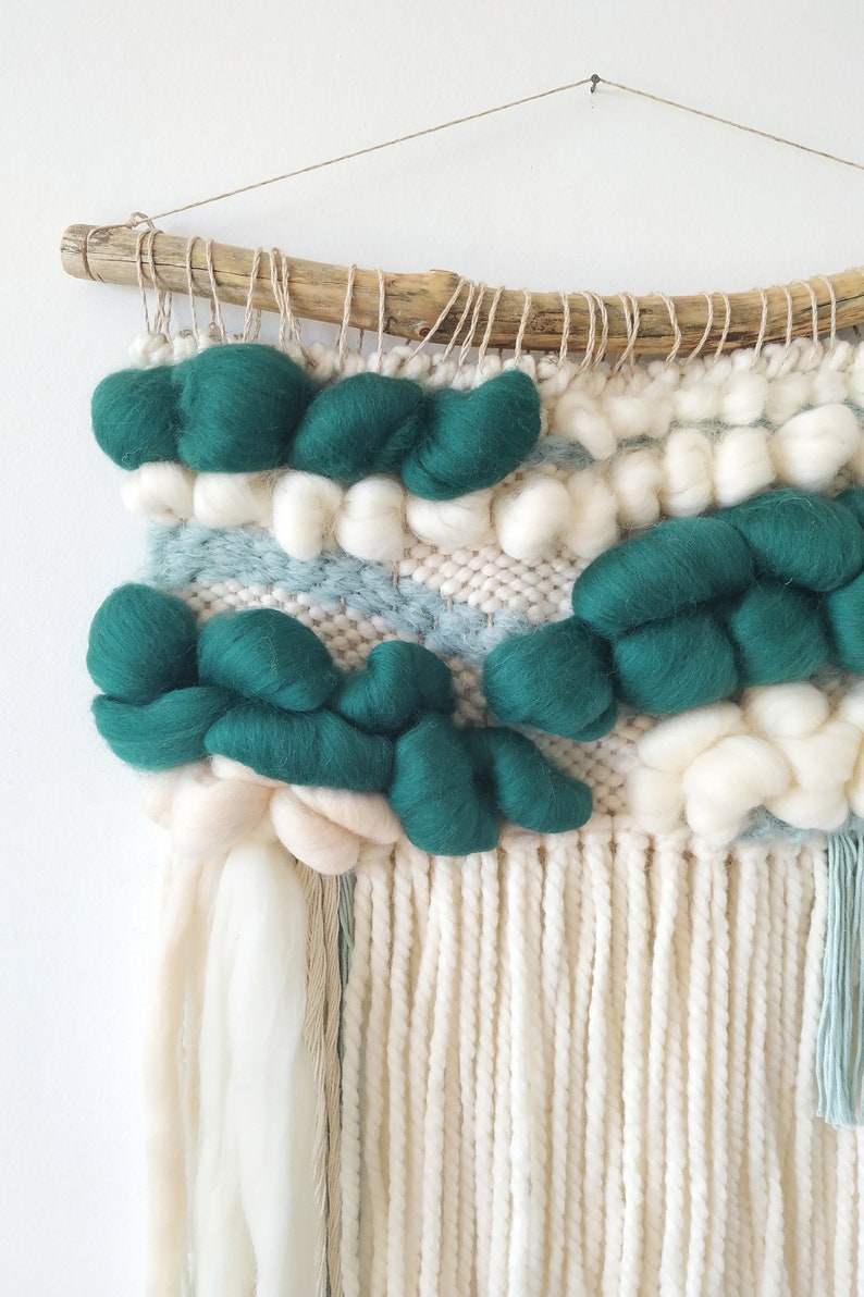May include: A woven wall hanging with a driftwood frame. The hanging features a green and white color scheme with a variety of textures. The hanging has a fringe of white yarn.
