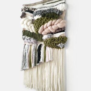 Woven wall hanging with an earthy, natural aesthetic. The wall hanging features a combination of olive green and beige tones, with textured, intricate weaving patterns. The design includes a central circular motif with hanging fringe details at the bottom, creating a bohemian and rustic look.