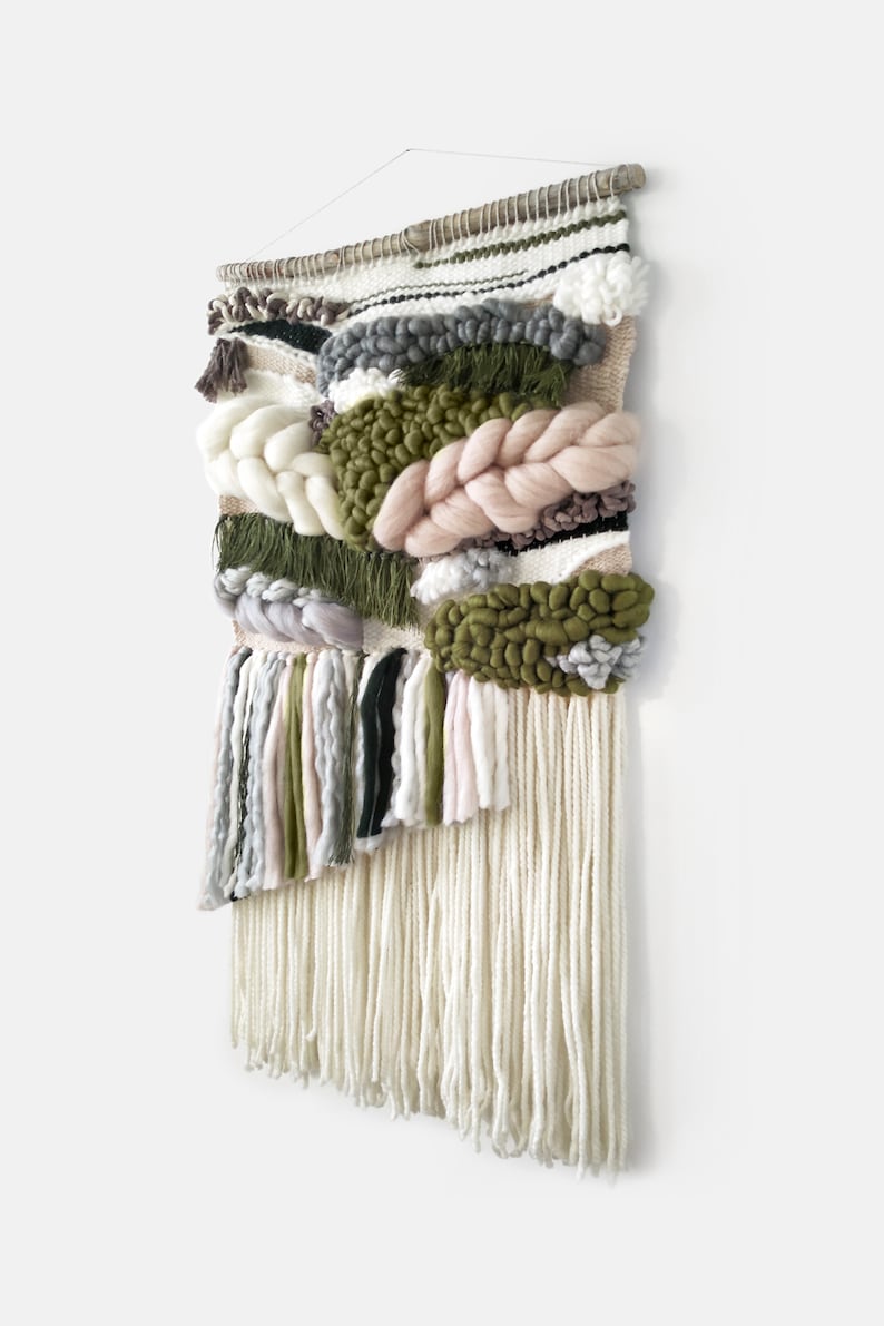Woven wall hanging with an earthy, natural aesthetic. The wall hanging features a combination of olive green and beige tones, with textured, intricate weaving patterns. The design includes a central circular motif with hanging fringe details at the bottom, creating a bohemian and rustic look.