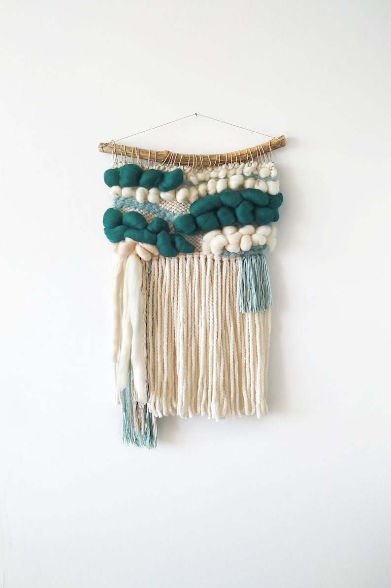 A small woven wall hanging in teal blue with intricate patterns and fringed edges, displayed against a plain background.