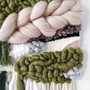 Detail of a woven wall hanging with an earthy, natural aesthetic. The wall hanging features a combination of olive green and beige tones, with textured, intricate weaving patterns. The design includes a central circular motif with hanging fringe details at the bottom, creating a bohemian and rustic look.