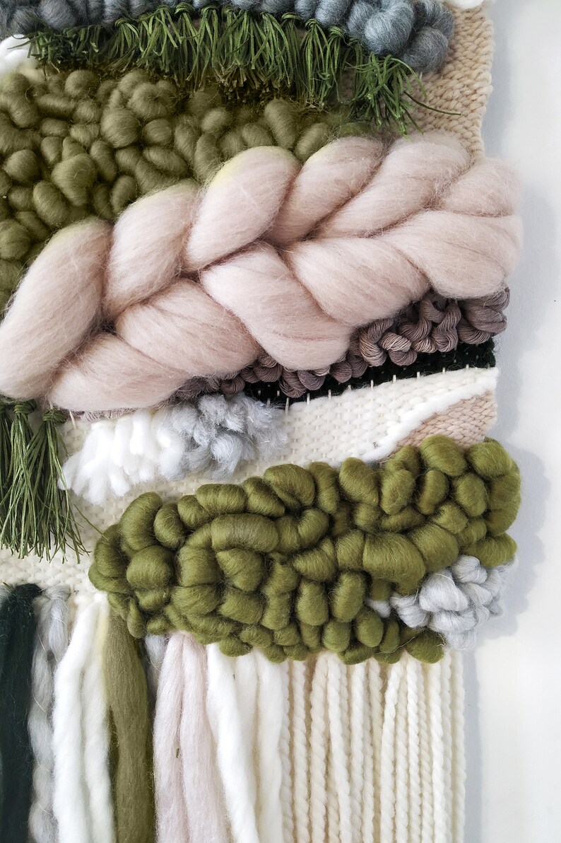 Detail of a woven wall hanging with an earthy, natural aesthetic. The wall hanging features a combination of olive green and beige tones, with textured, intricate weaving patterns. The design includes a central circular motif with hanging fringe details at the bottom, creating a bohemian and rustic look.
