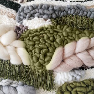 Detail of a woven wall hanging with an earthy, natural aesthetic. The wall hanging features a combination of olive green and beige tones, with textured, intricate weaving patterns. The design includes a central circular motif with hanging fringe details at the bottom, creating a bohemian and rustic look.