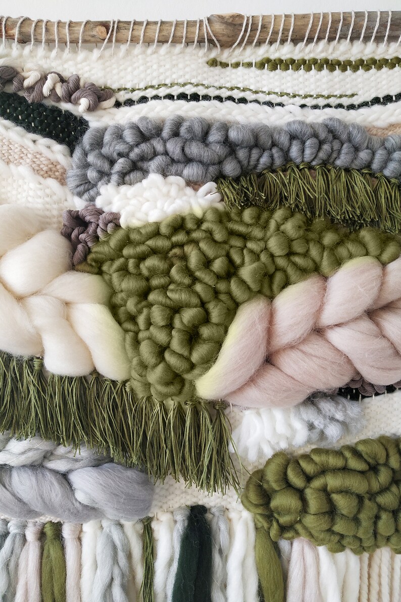 Detail of a woven wall hanging with an earthy, natural aesthetic. The wall hanging features a combination of olive green and beige tones, with textured, intricate weaving patterns. The design includes a central circular motif with hanging fringe details at the bottom, creating a bohemian and rustic look.