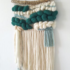 A small woven wall hanging in teal blue with intricate patterns and fringed edges, displayed against a plain background.