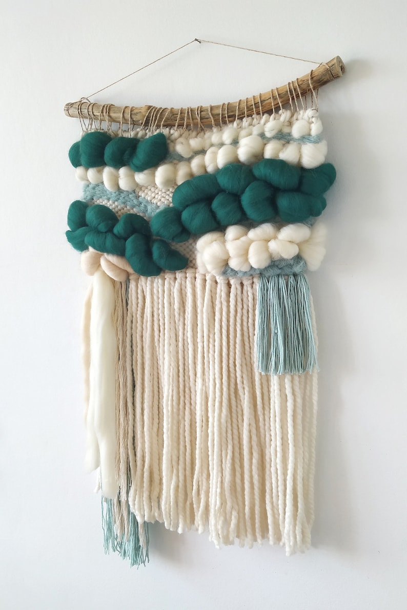 A small woven wall hanging in teal blue with intricate patterns and fringed edges, displayed against a plain background.