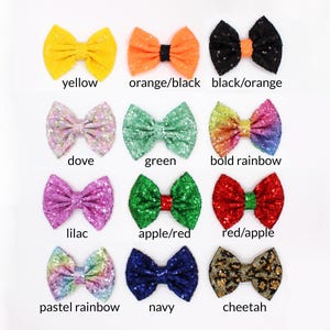 May include: A collection of twelve sequined hair bows arranged in a grid. Colors include yellow, orange/black, black/orange, dove, green, bold rainbow, lilac, apple/red, red/apple, pastel rainbow, navy, and cheetah print, each with a center knot.