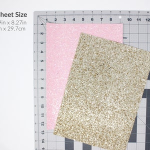 May include: Two sheets of glitter paper, one pink and one gold, are laid out on a cutting mat. The mat has inch and centimetre markings. The text "A4 Sheet Size" is printed in black at the top left corner of the image. The text "11.69in x 8.27in" and "21cm x 29.7cm" is printed below the text "A4 Sheet Size".