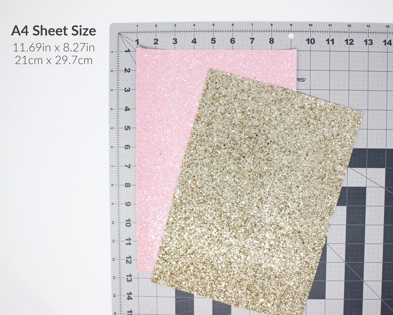 May include: Two sheets of glitter paper, one pink and one gold, are laid out on a cutting mat. The mat has inch and centimetre markings. The text "A4 Sheet Size" is printed in black at the top left corner of the image. The text "11.69in x 8.27in" and "21cm x 29.7cm" is printed below the text "A4 Sheet Size".
