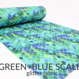 May include: A roll of glitter fabric with a green and blue scale pattern. The fabric is unrolled, revealing a repeating design of overlapping, semi-circular shapes in shades of green and blue. The text "GREEN+BLUE SCALES glitter fabric" is visible.