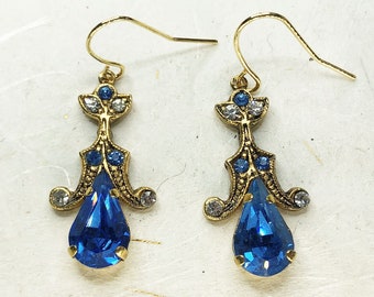Könnte beinhalten: Ein Paar goldfarbene Ohrhänger mit einem zentralen tropfenförmigen blauen Kristall, akzentuiert durch kleinere runde blaue und klare Kristalle in einem verzierten Metallrahmen mit Hakenverschluss für durchstochene Ohrläppchen.