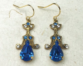 Könnte beinhalten: Ein Paar goldfarbene Ohrhänger mit einem großen, birnenförmigen blauen Kristall an der Unterseite. Der Metallrahmen hat eine verzierte, strukturierte Oberfläche und ist mit kleineren, runden blauen und klaren Kristallen in symmetrischer Anordnung.