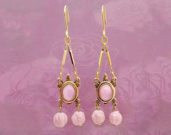 May include: A pair of gold-toned dangle earrings featuring a turtle-shaped centerpiece with an oval pink cabochon stone. Each earring hangs from a hook and includes two small, round, light pink carved beads suspended from the turtle's base.