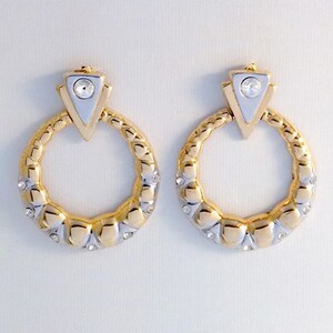 Può includere: Un paio di orecchini a cerchio color oro e argento con una finitura metallica testurizzata alternata. Ogni orecchino è sormontato da un elemento triangolare con un brillante rotondo trasparente e piccoli brillanti incastonati lungo il cerchio.