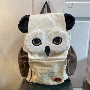 May include: A cream-colored backpack designed to resemble an owl. The backpack features brown suede-like side pockets and a front pocket with text. The owl's face has fluffy white accents, large yellow eyes, and black beak. The text on the front reads "MR. H. POTTER, The Cupboard under the Stairs, 4. Privet Drive, Little Whinging, SURREY."