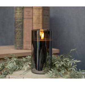 May include: A tall, cylindrical candle in a dark, reflective glass holder. The candle has a flickering, warm-toned flame. The holder has vertical ridges and sits on a surface with greenery and vintage books in the background. The candle is a decorative home accent.