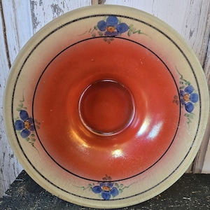 May include: A vintage, round bowl with a vibrant orange center and a cream-colored rim. The rim is decorated with blue floral accents and a black border. The bowl's design is symmetrical, with a circular indentation in the center.