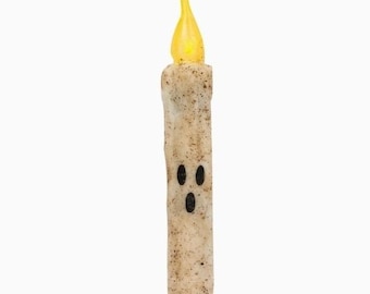 Puede incluir: Una vela cilíndrica con una superficie beige texturizada y moteada que presenta un rostro de fantasma negro impreso con dos ojos ovalados y una boca ovalada. La parte superior de la vela tiene forma de llama amarilla, imitando una luz.