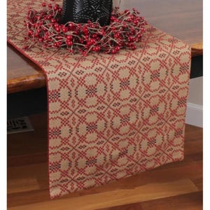 May include: A tan and red patterned table runner with a red border. A black candle holder and a red berry wreath sit on top of the runner. The runner has a geometric design.
