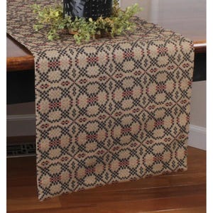 May include: A rectangular table runner with a repeating geometric pattern in black, red, and tan. The runner is draped over a wooden table, with a decorative black metal candle holder and greenery on top. The runner's design adds a rustic touch.