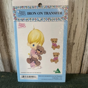 May include: A package of Precious Moments iron-on transfers. The package features a cartoon boy holding a teddy bear, with additional teddy bear designs. The text "IRON ON TRANSFER" is displayed at the top.
