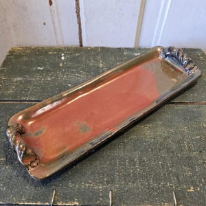 May include: A rectangular ceramic serving tray with a reddish-brown interior and a dark, metallic-looking rim. The tray has decorative rope-like handles at each end. The tray is approximately 12 inches long.
