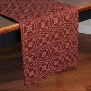May include: A red and beige patterned table runner. The runner features a repeating geometric design with a floral-like motif. The fabric is draped over a wooden table, showcasing the intricate pattern and color scheme.