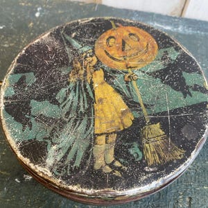 May include: Round vintage Halloween tin with a distressed finish. The lid features a child in a yellow dress and green witch's cloak holding a jack-o'-lantern and broom against a black background with green foliage.