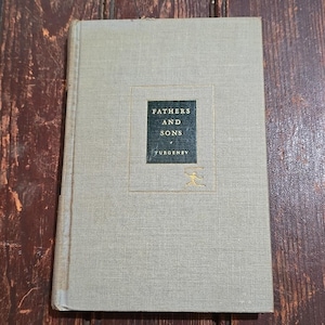 May include: A hardcover book titled "Fathers and Sons" by Turgenev. The book has a light gray cover with a dark green square containing the title and author's name. A gold illustration is below the author's name.