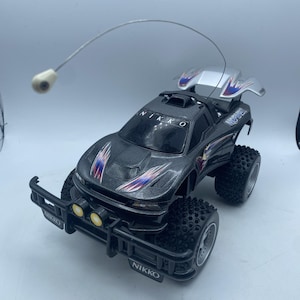 May include: A gray and black Nikko remote control car with large, knobby tires. The car has a silver spoiler and blue, red, and purple accents. The front bumper has two yellow lights and the word "NIKKO" is visible on the car.