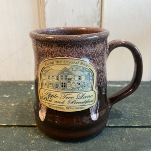 May include: A brown ceramic mug with a dark brown handle and rim. The mug features a cream-colored label with the text "Apple Tree Lane Bed and Breakfast Waupaca, Wisconsin" and an illustration of a house.