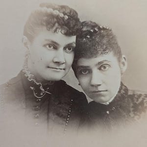 May include: A vintage black and white photograph featuring two women. They are close together, with their faces near each other. Both women are wearing dark clothing and have their hair styled up. The photograph has a soft focus.