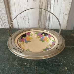 May include: A vintage serving bowl with a floral design in the center. The bowl has a silver-colored metal rim and handle. The floral design includes purple, orange, and blue flowers on a cream background.