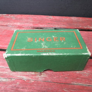 May include: A rectangular green cardboard box with orange trim and the word SINGER printed in orange on the top lid. The text MADE IN U.S.A. is printed in smaller orange letters below the brand name. The box shows signs of wear on the edges.
