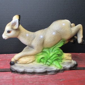 May include: A ceramic figurine of a leaping fawn with light tan fur, white spots, and dark-tipped ears. The fawn is positioned over a base of sculpted green grass and gray rocks, with its front legs tucked and hind legs extended in mid-air.