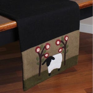 May include: Black table runner with a sheep and floral design. The runner features a black top portion and a lower section with a sheep, trees, and flowers in shades of brown, green, and red. The runner is made of a soft material.