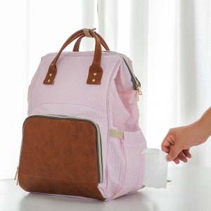 May include: A pink and white striped diaper bag with a brown faux leather front pocket. The bag has a zippered pocket on the side and a zippered top closure.